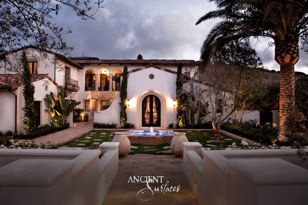 Estate at dusk with a Kronos limestone forecourt, a carved stone fountain, and lit arcades