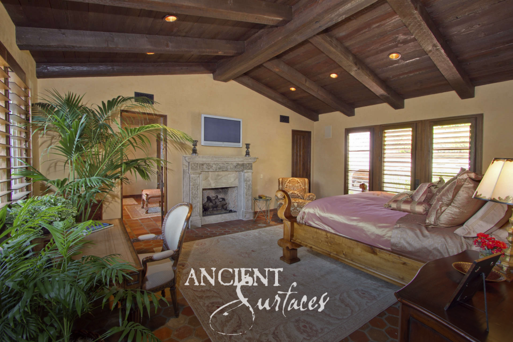 Bedroom beneath a dark timber beamed ceiling with a limestone mantel light in tone and ornate in detail above terracotta floors