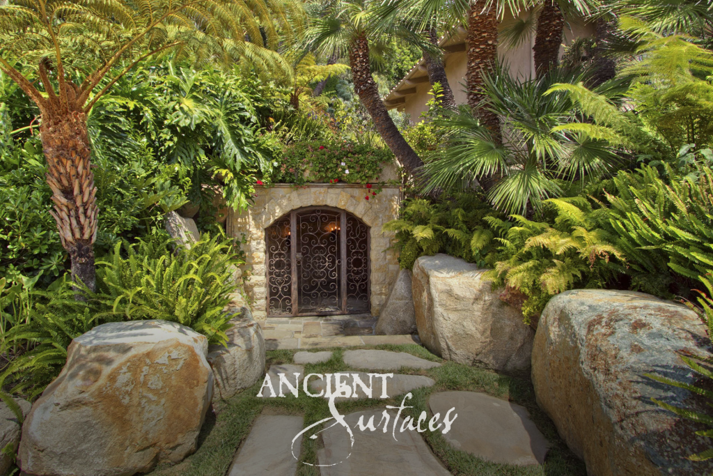 Garden pathway of irregular edged limestone slabs running through ferns and palms to an arched stone entry with a wrought iron gate